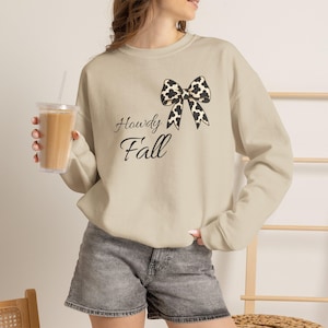Könnte beinhalten: Ein beigefarbenes Sweatshirt mit Rundhalsausschnitt, verziert mit einer schwarz-weißen Schleifengrafik im Kuhfellmuster über dem schwarzen kursiven Schriftzug Howdy Fall. Das Kleidungsstück hat lange Ärmel sowie gerippte Bündchen an Ausschnitt, Ärmeln und Saum.