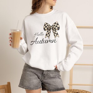 Könnte beinhalten: Ein weißes Langarm-Sweatshirt mit einer schwarz-weiß karierten Schleifengrafik über dem schwarzen kursiven Text "Hello Autumn". Das Kleidungsstück wird mit grauen Jeansshorts kombiniert und ein durchsichtiger Plastikbecher mit einem beigen Getränk wird gehalten.