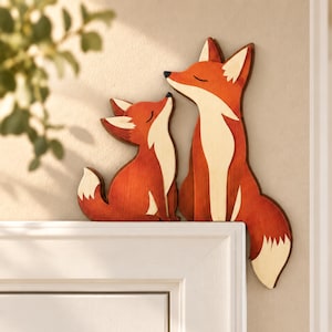 May include: Two wooden fox cutouts in orange and cream colors, designed to sit on a corner edge. The larger fox sits upright while the smaller fox sits beside it, both featuring closed eyes and pointed ears with white-tipped tails.