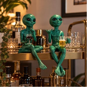 Puede incluir: Dos figuras de alienígenas verdes sentadas en un estante. El alienígena de la izquierda sostiene una copa de vino y hace un gesto de silencio. El de la derecha hace un gesto de shaka mientras sostiene una cerveza. Tienen grandes ojos negros.