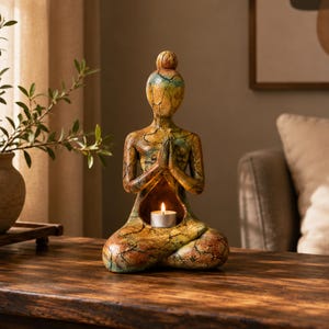 Peut inclure: Une sculpture de personnage assis en pose méditative, les mains jointes en prière. La surface présente une finition texturée et craquelée dans des tons jaune, orange et bleu sarcelle. Une cavité dans le torse contient une petite bougie chauffe-plat blanche allumée.