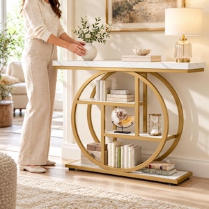 May include: A gold metal console table with a white marble-patterned top and two circular side frames. The table features three interior shelves holding books, a decorative nautilus shell, and an hourglass. A lamp sits on the top surface.
