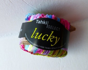 May include: A skein of Tahki Select Lucky yarn in a multi-colored striped pattern. The yarn is made in Italy exclusively for Tahki. The label reads "Tahki Select Lucky" and "Made in Italy Exclusively for Tahki - Stacy Charles Inc."