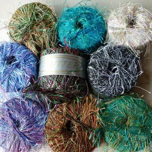 May include: A collection of colorful yarn balls in shades of blue, green, purple, and brown. The yarn has a textured, fringed appearance, with some balls wrapped with a label that reads "Glitter". Ideal for crafting and textile projects.