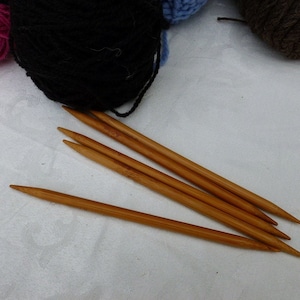 May include: A collection of knitting supplies, including several wooden knitting needles and balls of yarn in pink, black, blue, and brown. The needles are a light brown color and are pointed at one end.