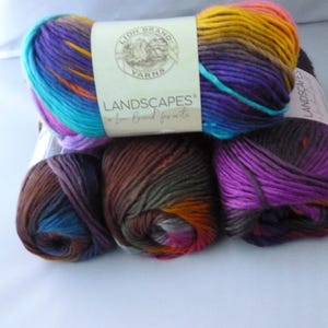 May include: A collection of colorful yarn skeins. The yarn is a blend of blues, purples, oranges, and yellows. The label reads "LANDSCAPES a Lion Brand favorite". The yarn is likely used for knitting or crocheting projects.