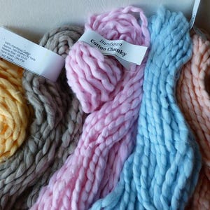 May include: A collection of handspun cotton chunky yarn in various colors. The yarn is thick and textured, with shades of yellow, gray, pink, blue, and peach. Each skein is labeled with details about the yarn's composition and care instructions.