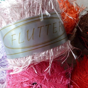 May include: Close-up of colorful yarn skeins in shades of pink, orange, purple, and brown. A label reading "FLUTTER" is wrapped around a pink skein. The yarn has a fuzzy, textured appearance, ideal for crafting projects.