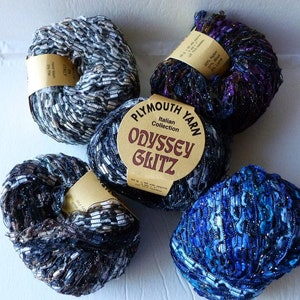 May include: Four skeins of yarn in different colors, including black, white, blue, and purple. The yarn is a sparkly, textured type. The label on one skein reads "Plymouth Yarn Italian Collection Odyssey Glitz".