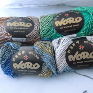 May include: Four skeins of Noro yarn in various colorways, including brown, green, blue, and grey tones. Each skein features a black paper label with the text Noro, the world of nature, and Made in Japan, wrapped around the center of the yarn.
