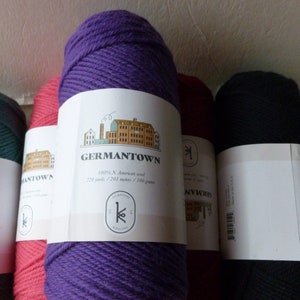 May include: Close-up of several skeins of Germantown wool yarn in various colors. The colors include teal, pink, purple, red, and black. Each skein has a white label with the Germantown Woolen logo and product information.