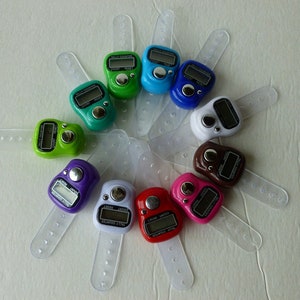 May include: A set of ten colorful digital tally counters with clear plastic wristbands. Each counter has a button to increment the count and a digital display showing the current count. The counters are arranged in a circle on a white surface.
