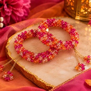 Op de afbeelding: Twee ronde armbanden gemaakt van kleine, gefacetteerde kralen in levendige tinten roze, oranje en helder wit. Elke armband heeft fijne goudkleurige kettinkjes die aan de zijkant hangen en eindigen in trosjes van bijpassende veelkleurige kralen.