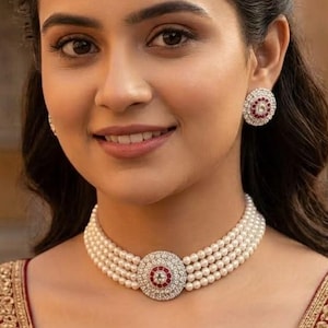 May include: A multi-strand pearl choker necklace featuring a central circular pendant with a red stone surrounded by small white crystals. Matching circular stud earrings with a red stone and white crystal border are worn with a red embroidered garment.