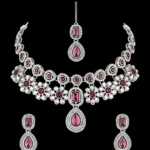 Puede incluir: Un conjunto de joyería en tono plateado que incluye un collar, un par de pendientes y un maang tikka. Cada pieza está adornada con piedras de color rosa intenso rectangulares y en forma de lágrima, rodeadas de pequeños cristales claros.