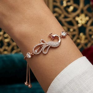 May include: A rose gold-toned bracelet featuring a peacock-shaped centerpiece encrusted with small clear stones and a single red stone for the eye. The design includes two larger clear stones on the chain and an adjustable sliding clasp mechanism.