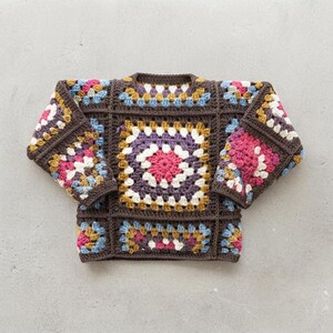May include: A long-sleeve sweater constructed from multiple crocheted granny squares. The garment features a dark brown base color with accent squares in shades of bright pink, white, mustard yellow, and light blue arranged in a geometric pattern.