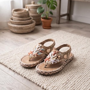 May include: A pair of tan woven straw sandals featuring a thong-style strap adorned with decorative pastel-colored starfish and coral motifs. The sandals have a slingback strap and a braided edge detail around the sole, providing a textured finish.