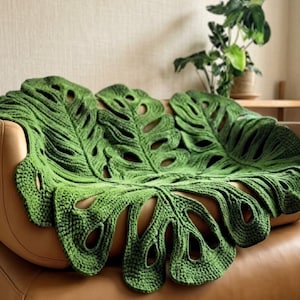 Può includere: Una grande coperta all'uncinetto verde a forma di foglia di monstera con ritagli e venature caratteristiche. Il tessuto presenta un aspetto strutturato e cucito a mano con filato verde scuro, drappeggiato su un divano in pelle color cuoio.