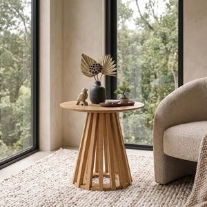 May include: A round light-toned wooden side table with a slatted base. On the table sits a black vase containing dried palm leaves and a lotus pod, a small carved wooden bird figurine, and a miniature bonsai tree on a small wooden tray.