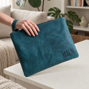 Könnte beinhalten: Eine petrolfarbene Leder-Clutch mit Handschlaufe und den geprägten Buchstaben MKN in der unteren rechten Ecke. Die rechteckige Tasche hat eine glatte, matte Oberfläche mit subtilen natürlichen Variationen in der tiefblau-grünen Textur.