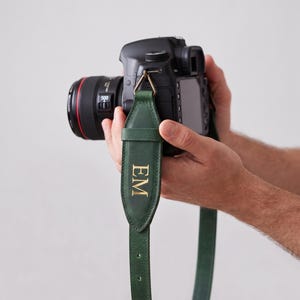 Op de afbeelding: Een donkergroene lederen camerariem bevestigd aan een zwarte digitale camera. De riem heeft een breder gewatteerd gedeelte met de goudkleurige in reliëf gedrukte initialen EM verticaal gecentreerd, vastgezet met een metalen driehoekige ring.
