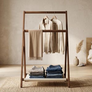 Puede incluir: Un perchero de madera en forma de A con una barra horizontal para colgar prendas y un estante inferior. El perchero sostiene una bufanda beige, una camisa clara, una camiseta de manga larga en perchas y dos pilas de pantalones vaqueros doblados.