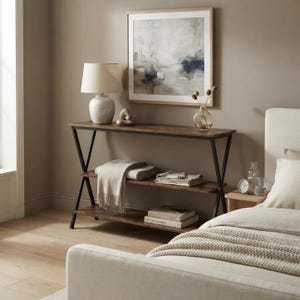 Peut inclure: Une console avec une finition en bois foncé et une base en métal noir en forme de X. La table comporte deux étagères inférieures contenant des textiles pliés et des livres. Une lampe avec un abat-jour beige, un vase en verre et un tableau abstrait.