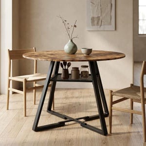 Peut inclure: Une table à manger ronde avec un plateau en bois brun et un cadre en métal noir doté d'une étagère de rangement inférieure en maille. La table est accompagnée de deux chaises en bois clair avec des assises tressées. Un vase et des bocaux.