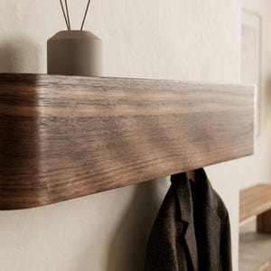 Peut inclure: Une étagère murale flottante en bois brun foncé avec un bord gauche arrondi et lisse. L'étagère présente un motif de grain horizontal et une surface supérieure plate. Un manteau gris foncé est suspendu sous l'étagère fixée au mur.