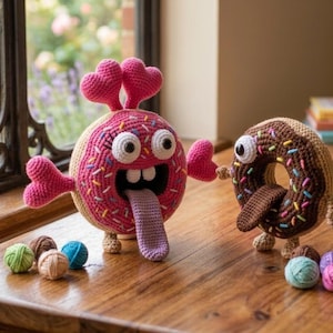Könnte beinhalten: Zwei gehäkelte Donut-Kreaturen mit großen weißen Augen, breiten Mündern und langen Zungen. Der größere Donut ist rosa mit bunten Streuseln und herzförmigen Antennen, während der kleinere braun mit Schokoladenglasur und Streuseln ist.