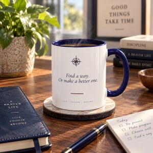 Peut inclure: Un mug en céramique blanche avec un intérieur et une anse bleu foncé. L'avant présente un graphique de boussole, le texte "Find a way. Or make a better one.", une ligne rouge horizontale et les mots "LIVE CHOOSE BECOME" en petites lettres.