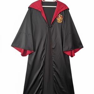 May include: Black robe with a red-lined hood and sleeves, featuring a Gryffindor crest. The robe is hanging on a white hanger against a white background. The robe has a button closure.