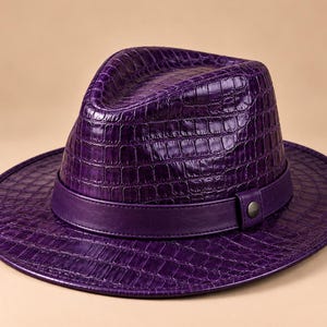 Op de afbeelding: Een dieppaarse fedora-hoed met een textuur van krokodillenleer over de kroon en de brede rand. De hoed is geaccentueerd met een gladde, effen paarse band rond de basis, die is vastgezet met een kleine, ronde metalen knop.