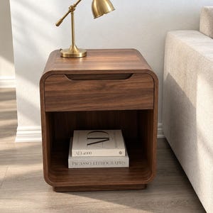 May include: A dark wood nightstand with rounded corners, featuring a single top drawer with a recessed handle and an open lower shelf. A brass-toned metal desk lamp sits on top, and two books titled AD at 100 and Picasso Lithographs are on the shelf.
