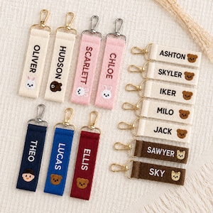 May include: A collection of rectangular fabric keychains featuring embroidered names and small animal icons. The keychains come in various colors including cream, pink, navy, blue, red, and brown, each attached to a gold-toned metal lobster clasp.