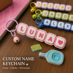 May include: A custom name keychain featuring the name LUNA in pink letters on white square blocks with a light purple ring and a small pink bell. Additional keychains with names DREAM, JAYDE, and ALEXI are visible, along with a blue paddle piece.