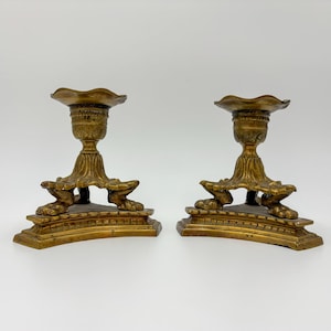 May include: A pair of matching brass candle holders featuring a scalloped top rim, a central stem with vertical fluting, and a base supported by three distinct claw-style feet resting on a curved, tiered platform with decorative edge detailing.