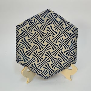May include: A hexagonal ceramic tile featuring a repeating dark blue and cream-colored woven pattern. The tile is displayed upright on a small, light-colored plastic stand, showcasing the intricate geometric design across its flat surface.