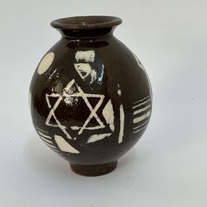 May include: A round, dark brown ceramic vase with a glossy finish. The surface is decorated with white, hand-painted geometric patterns, including a prominent six-pointed star, various abstract shapes, and sets of parallel lines around the body.