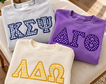 Könnte beinhalten: Drei Rundhals-Sweatshirts mit griechischen Buchstaben. Das graue Sweatshirt hat marineblaue Vichy-Karo-Buchstaben KΣΨ. Das lila Sweatshirt hat weiß gepunktete Buchstaben ΔΓΘ. Das cremefarbene Sweatshirt hat gelbe Vichy-Karo-Buchstaben ΛΔΩ.
