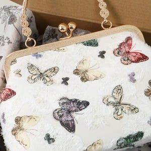 May include: A white clutch bag featuring a multi-colored butterfly print and a textured, frayed fabric overlay. The bag has a gold-toned metal frame, a kiss-lock closure, and a decorative gold-toned metal handle with circular floral accents.