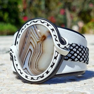 Puede incluir: Una pulsera de brazalete ancho con una gran piedra de ágata ovalada de bandas marrones y blancas. La piedra está enmarcada por detalles de cuero negro y blanco, con una banda de cuero blanco y un patrón texturizado de piel de serpiente.