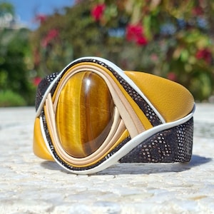 Puede incluir: Una pulsera de brazalete ancha que presenta una gran piedra preciosa ovalada de ojo de tigre marrón y dorado en el centro. La pulsera está construida con paneles de cuero en capas en amarillo mostaza, beige y un material texturizado marrón oscuro.