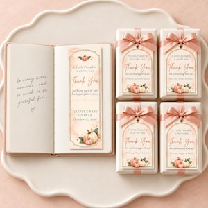 Könnte beinhalten: Ein offenes Notizbuch mit einem Lesezeichen, das Kürbisillustrationen und den Text A Little Pumpkin is on the way zeigt, neben vier rechteckigen Geschenkboxen mit rosa Schleifen und passenden Dankesanhängern.