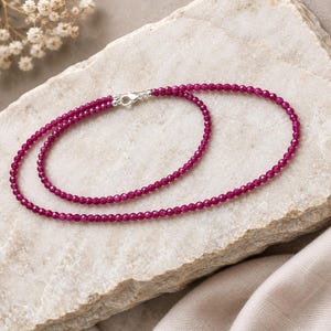 May include: A delicate necklace composed of small, faceted, deep magenta-colored beads arranged in a circular pattern. The necklace features a polished silver-toned lobster claw clasp that secures the two ends of the beaded strand together.
