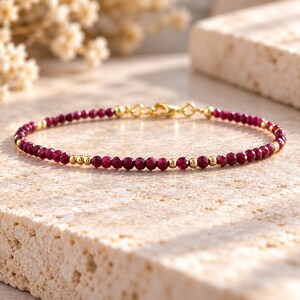 May include: A delicate bracelet featuring small, faceted deep red gemstone beads interspersed with tiny, polished gold-tone metal spheres. The piece is finished with a gold-tone lobster claw clasp and matching jump rings for a secure closure.