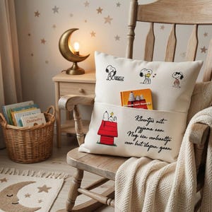 May include: A cream-coloured throw pillow with a front pocket featuring Snoopy illustrations and Dutch text. The top section shows Snoopy in various poses, while the pocket displays a red doghouse and the text: Kleentjes uit, pyjama aan. Nog één verhaaltje voor het slapen gaan.