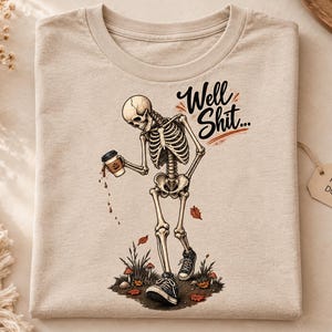 Puede incluir: Una camiseta beige con el gráfico de un esqueleto caminando mientras derrama una taza de café. El esqueleto lleva zapatillas negras y está rodeado de pequeñas setas y hojas de otoño. El texto "Well Shit..." está impreso en letra cursiva negra.