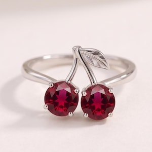 Peut inclure: Une bague argentée ornée de deux pierres rondes à facettes rouge foncé disposées pour ressembler à des cerises. Le design comprend une petite feuille argentée détaillée fixée aux tiges, avec des pierres maintenues par quatre griffes.